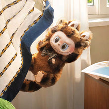 Load image into Gallery viewer, A plush monkey doll with large blue eyes, a patterned necktie, and brown-and-black striped fur, posed hanging beside a striped blanket in a bright indoor room.