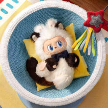 Load image into Gallery viewer, A plush toy character with fluffy white fur, dark brown ears and paws, and blue eyes, posed inside a round woven basket on a yellow pillow. A red star-shaped hanging toy is visible to the right.