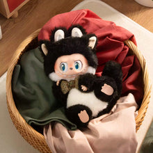 Load image into Gallery viewer, A black-and-white plush character doll with blue eyes and a small green bow tie, nestled in a woven basket with folded fabric in the background.
