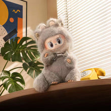 Load image into Gallery viewer, A plush toy character with large eyes, wearing a fuzzy gray animal-like hood, sitting on a wooden surface in a styled room with blinds, a plant, and a yellow rotary phone.
