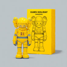 Load image into Gallery viewer, A yellow KAWS: Holiday collectible figure stands in front of its matching yellow packaging box on a light gray background. The box features a black line art illustration of the figure and the text 'KAWS:HOLIDAY HANGZHOU'.

