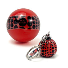 Load image into Gallery viewer, A red spherical keychain accessory with black polka dots and a silver metal key ring, shown alongside a larger matching red sphere on a white background. The smaller sphere has the text 'YAYOI KUSAMA' printed on it.
