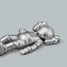 Load image into Gallery viewer, A metallic silver toy figure of a sleeping astronaut lying on its side against a solid gray background.

